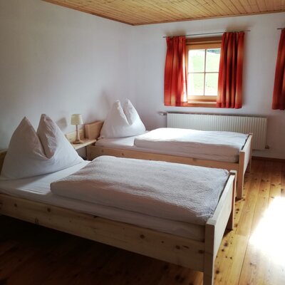 A bedroom in the farmhouse featuring two single beds, wooden flooring, a wooden ceiling, and a window with red curtains.