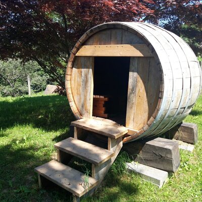 Outdoor barrel sauna with wooden steps on the farmhouse grounds.