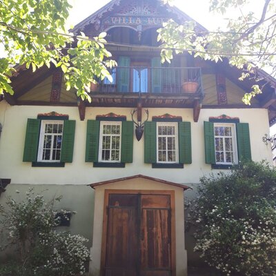 The exterior of the farmhouse, featuring traditional gabled architecture, green shutters, an upper-floor balcony, and a large wooden entrance.
