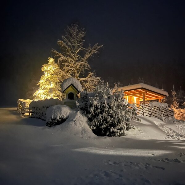 The farmhouse exterior illuminated at night, surrounded by snow-covered trees and a warm glow from the covered porch.