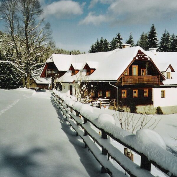 The Farm House exterior in winter, featuring snow-covered roofs, balconies, and a surrounding forest.