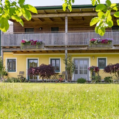 Exterior view of the farmhouse with a yellow facade, balcony with flower boxes, and ground-floor access to the double rooms. (c) Elisabeth Fröhlich Photography