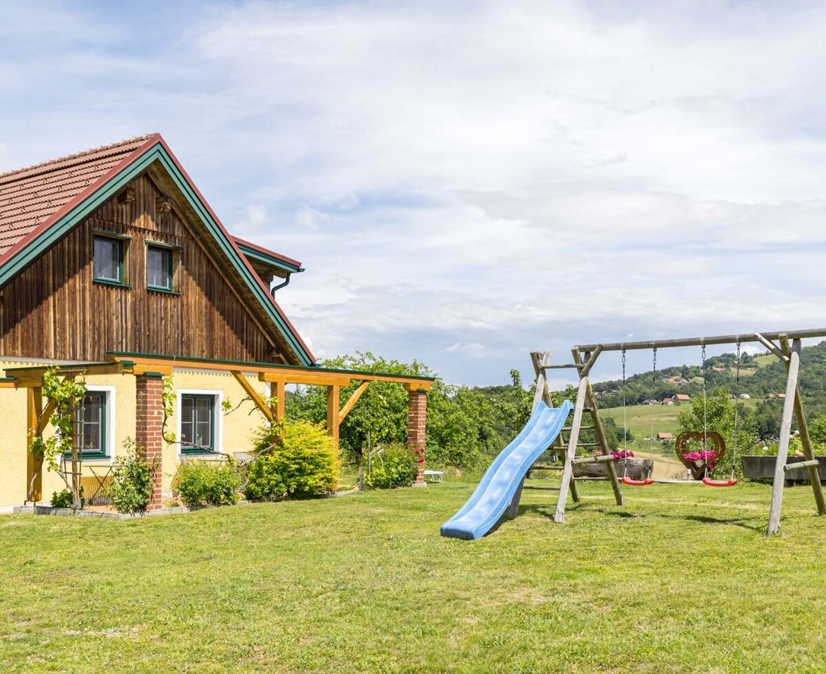 The exterior of the farmhouse with a lawn and a children's playground including a slide and swings. (c) Elisabeth Fröhlich Photography