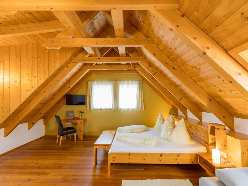 Spacious double room in the attic of the farmhouse with wooden beams, wooden floor, double bed, desk, and TV. (c) Elisabeth Fröhlich Photography