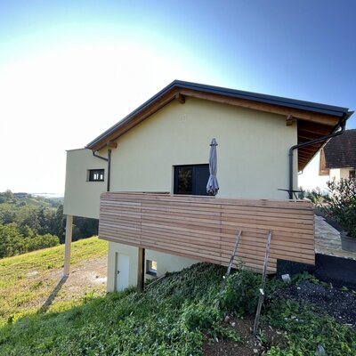 The farmhouse features an exterior wooden balcony with an umbrella, overlooking the green landscape.
