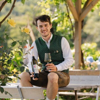 A man in traditional attire holds a wine bottle and glass under a vine-covered pergola at the Haus.