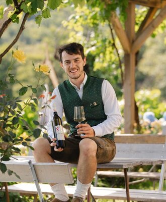 A man in traditional attire holds a wine bottle and glass under a vine-covered pergola at the Haus.