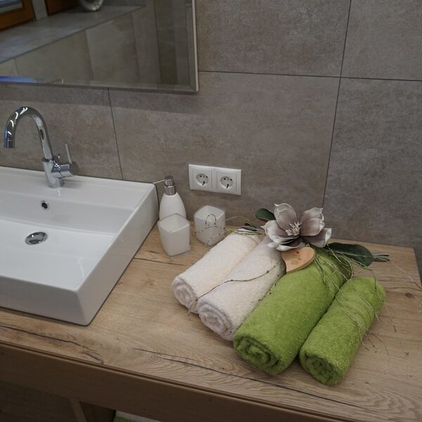 The Farm House bathroom features a white square sink with a chrome faucet, a wooden vanity top, a mirror above tiled walls, and readily available power outlets, complemented by guest towels and toiletries.