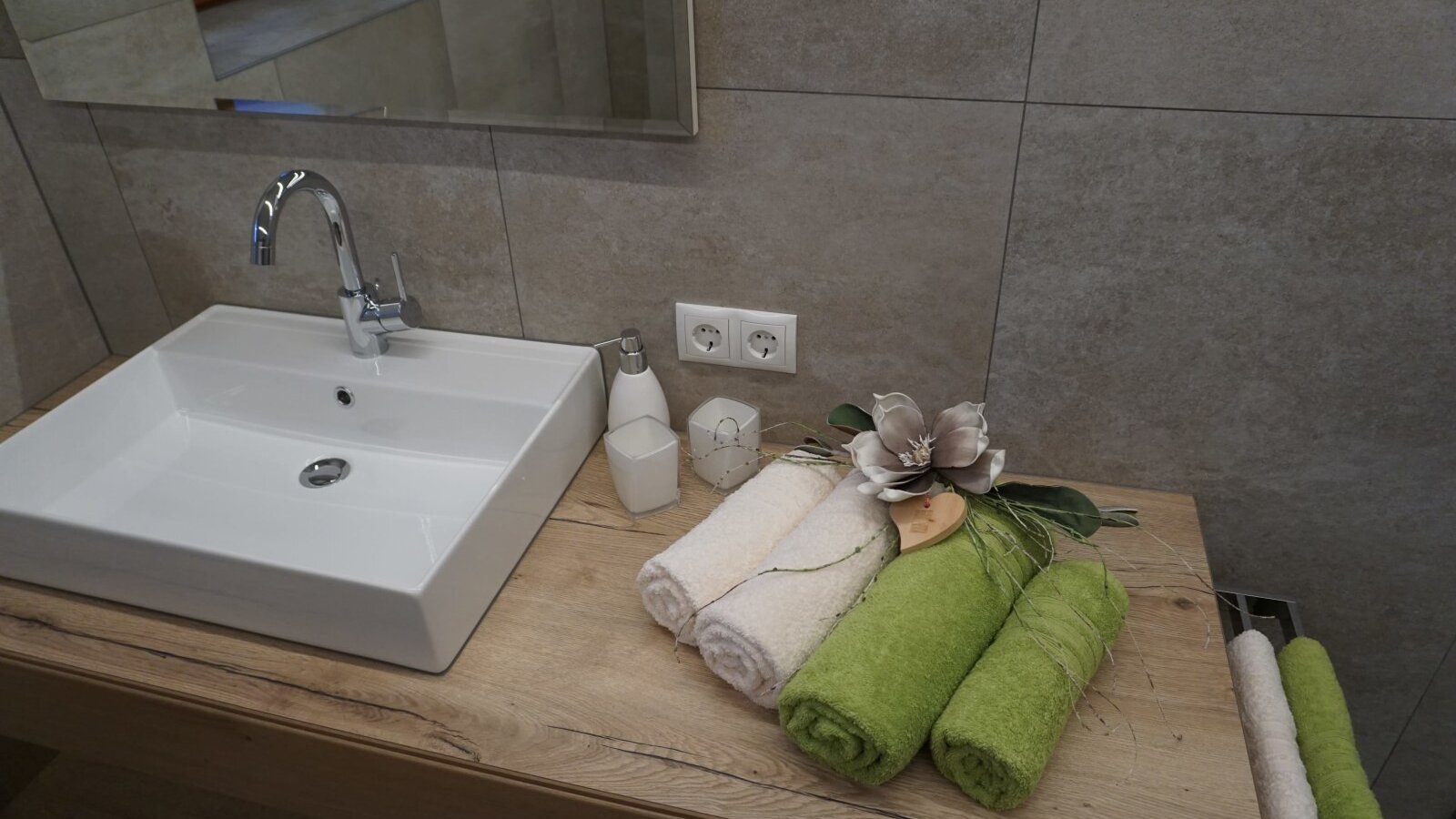 The Farm House bathroom features a white square sink with a chrome faucet, a wooden vanity top, a mirror above tiled walls, and readily available power outlets, complemented by guest towels and toiletries.