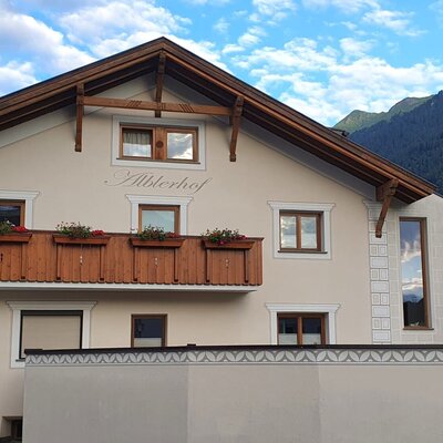 The exterior of the Alblerhof farmhouse, featuring traditional architecture, a wooden balcony with flower boxes, and a mountain view.