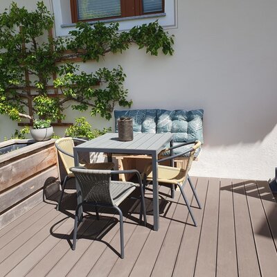 Farmhouse terrace with a dining table, four chairs, and a cushioned bench, surrounded by plants.