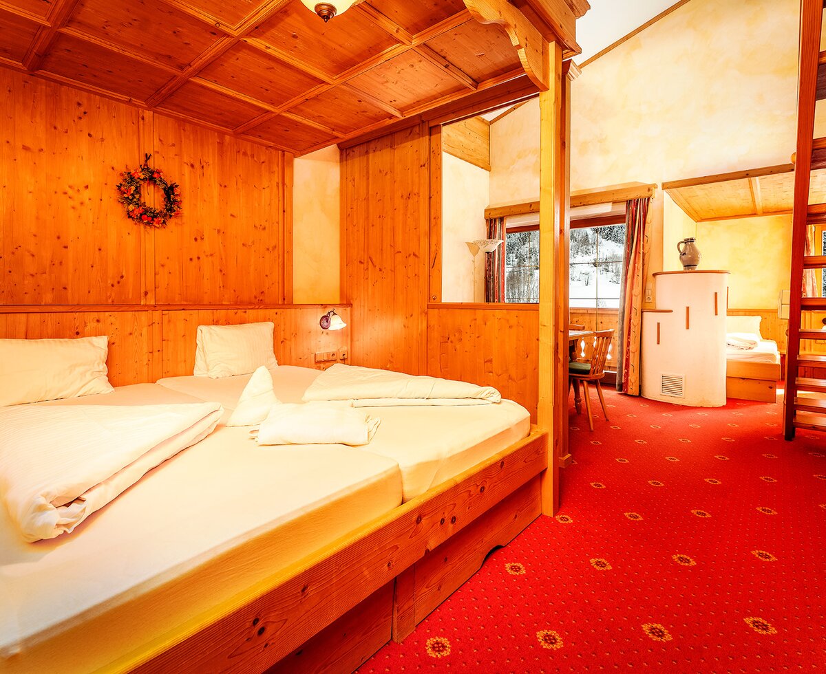 A suite in the farm house, featuring a wooden double bed, a separate sleeping area accessed by a ladder, a dining nook, a tiled stove, and views of a snowy landscape.