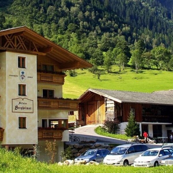 Exterior view of the farmhouse with balconies, a parking area, and a children's playground, set against a mountain backdrop.
