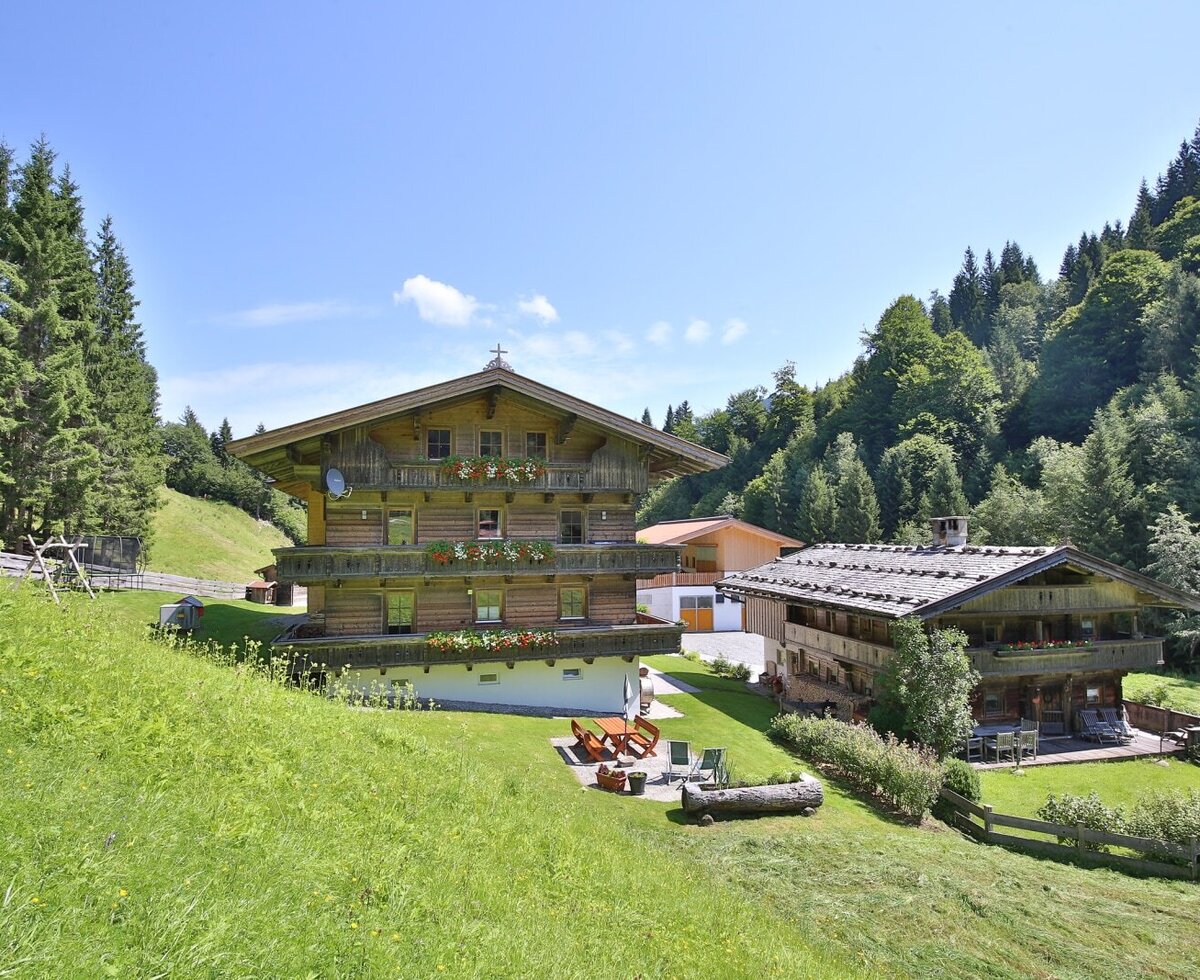 The farmhouse exterior, featuring traditional wooden construction, balconies with flower boxes, and an outdoor seating area on a grassy slope.