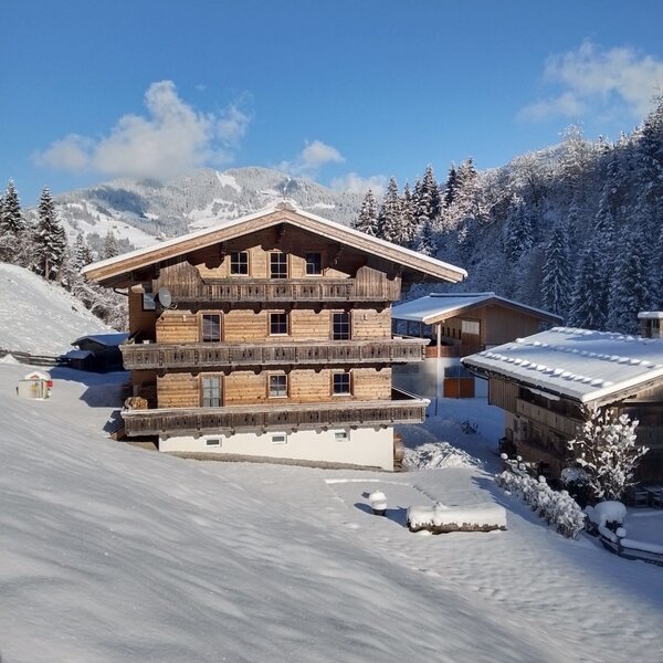 The farmhouse in a winter setting, with a wooden facade, balconies, and snow-covered mountains in the background.