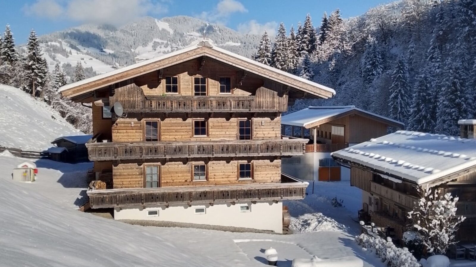 The farmhouse in a winter setting, with a wooden facade, balconies, and snow-covered mountains in the background.