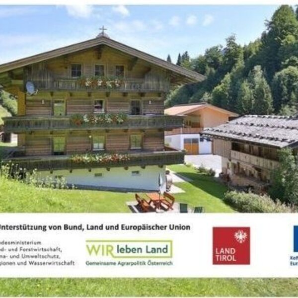 The inviting farmhouse surrounded by green mountain meadows, featuring traditional wooden balconies and a well-kept garden area.