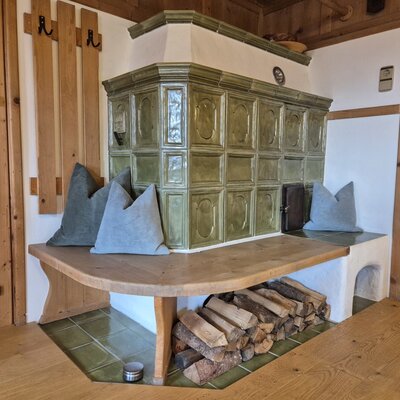 A traditional, green tiled stove in the farmhouse with a cozy wooden bench, soft pillows, and neatly stacked firewood.