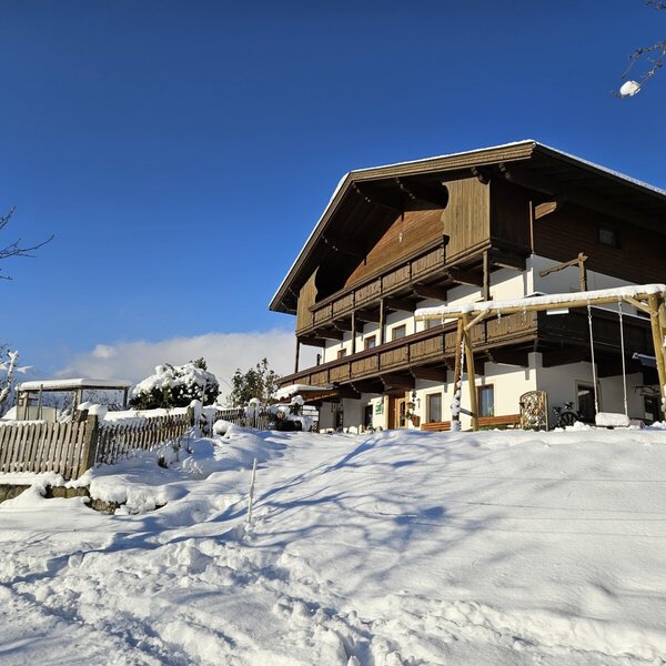 The Farm House exterior in winter, featuring wooden balconies, a children's playground, and snow-covered grounds under a clear blue sky.