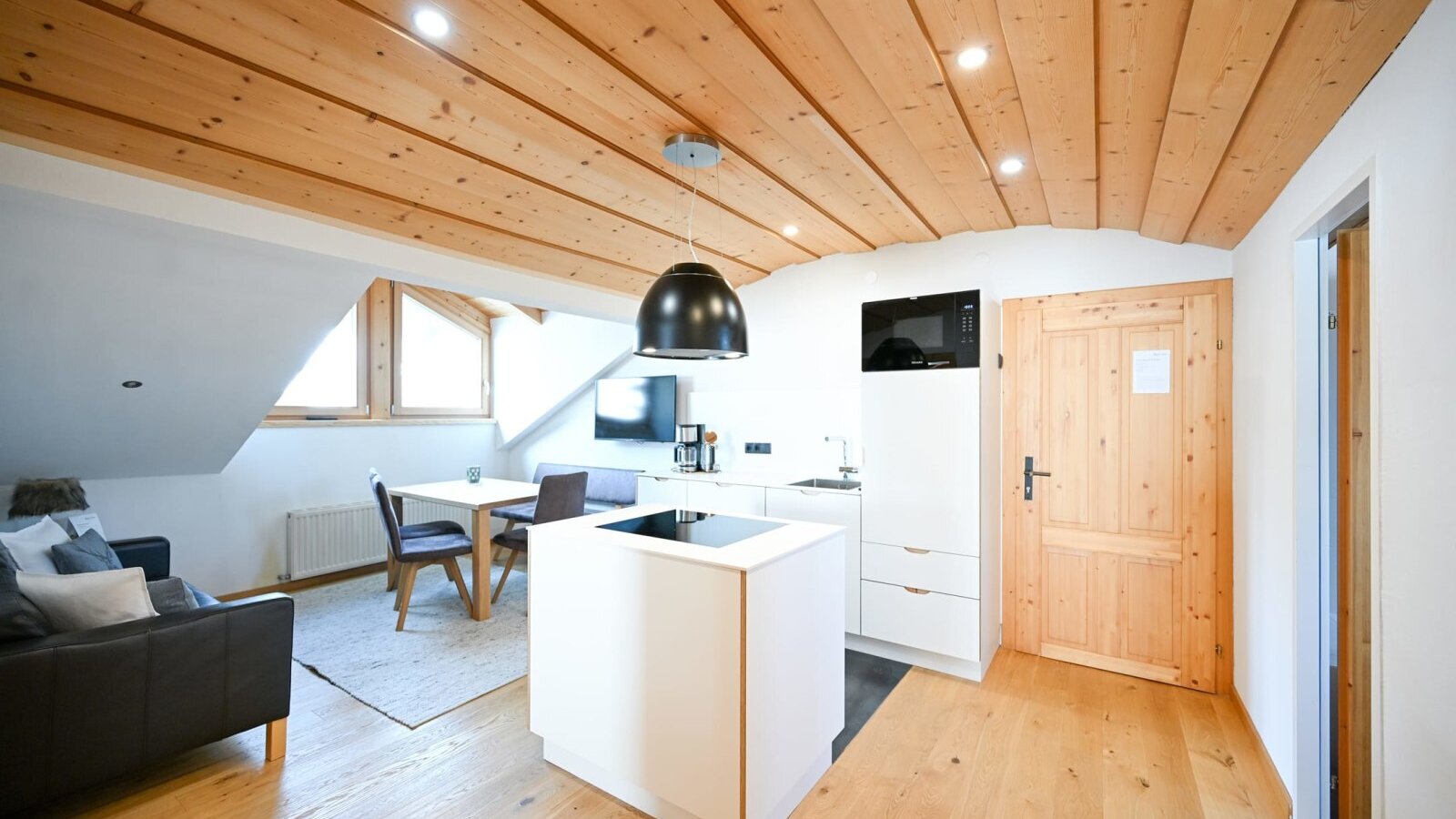 Open-plan living area of the apartment in the farmhouse, featuring a modern kitchen with an island, a dining area, a sofa, and a wooden ceiling.