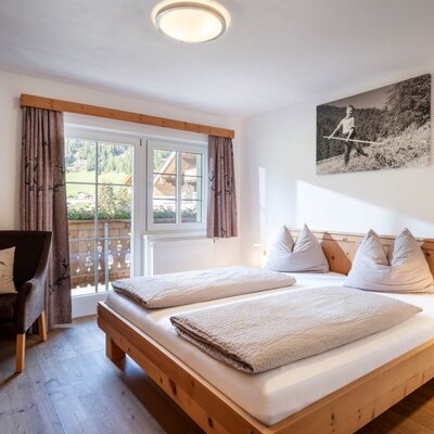 A cozy double bedroom at the farmhouse featuring warm wooden furniture, a comfortable armchair, and access to the balcony.