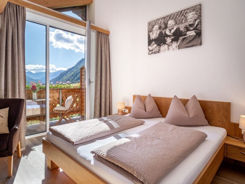 A cozy double room at the farmhouse with wooden furnishings, warm bedside lighting, and direct access to a sunny balcony with mountain views.