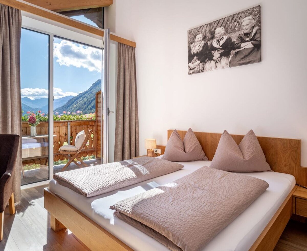 A cozy double room at the farmhouse with wooden furnishings, warm bedside lighting, and direct access to a sunny balcony with mountain views.
