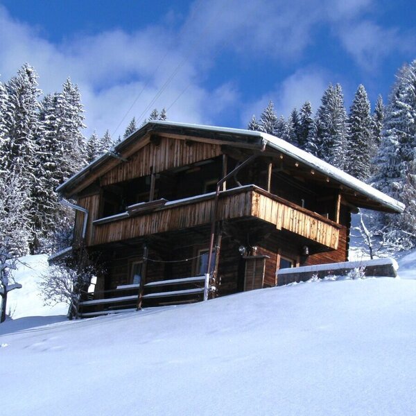 The wooden Alpine Hut exterior, featuring a balcony, surrounded by a snowy landscape with pine trees.