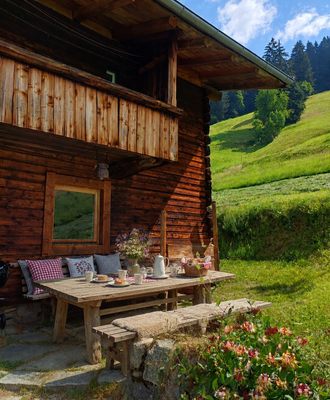 An inviting outdoor seating area at the Alm, featuring a rustic wooden table with cushions and views of the lush green hills.