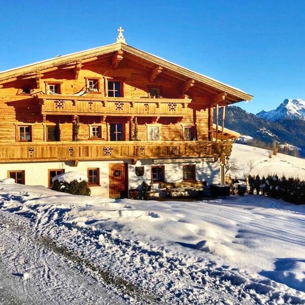 Exterior view of the wooden Farm House, featuring multiple balconies and surrounded by a snow-covered landscape with mountains in the background.