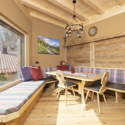 The dining area in the farmhouse features a wooden table with chairs and upholstered benches, a beamed wooden ceiling, and a large window with an outside view.