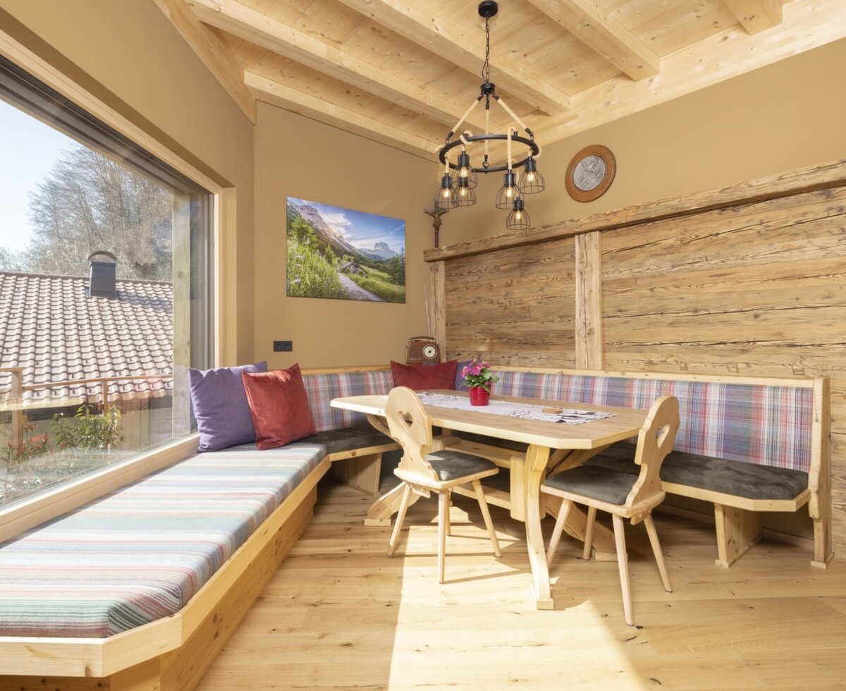 The dining area in the farmhouse features a wooden table with chairs and upholstered benches, a beamed wooden ceiling, and a large window with an outside view.