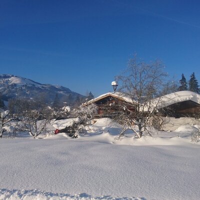 The farm house buildings with snow-covered roofs, set against a backdrop of snowy mountains and a clear blue sky.