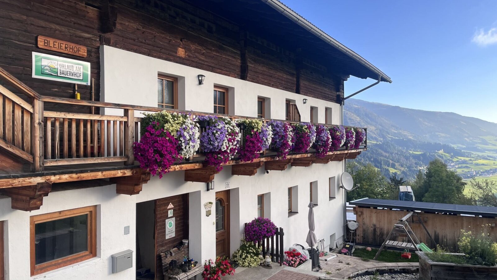 Exterior view of the farmhouse with a flower-decorated balcony, mountain view, and a children's swing in the garden.
