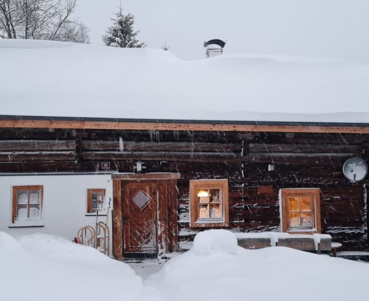 The Alpine Hut in winter, featuring a snow-covered exterior and a cleared path to the entrance amidst falling snow.