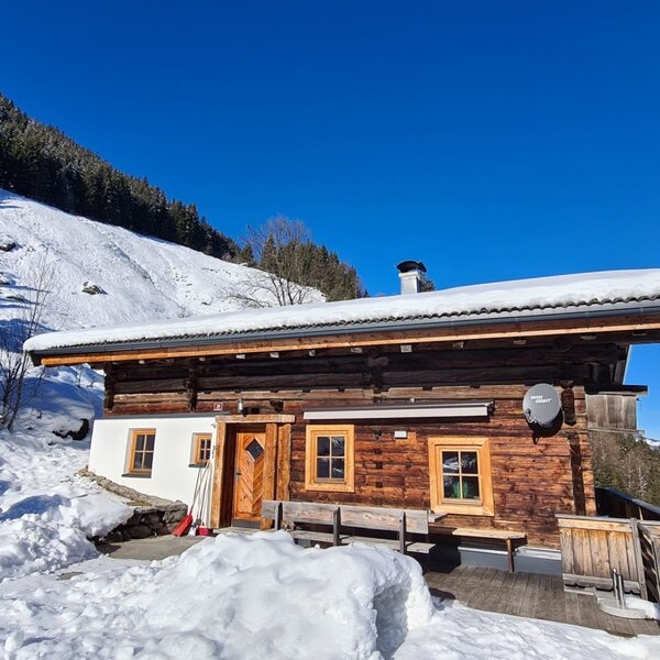 The Alm features a wooden facade and snow-covered roof, surrounded by snowy mountains under a blue sky, with an outdoor oven.