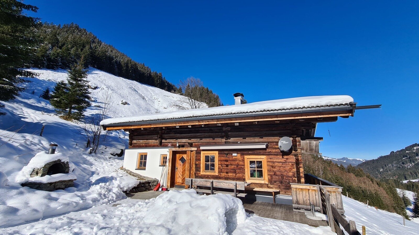 The Alm features a wooden facade and snow-covered roof, surrounded by snowy mountains under a blue sky, with an outdoor oven.