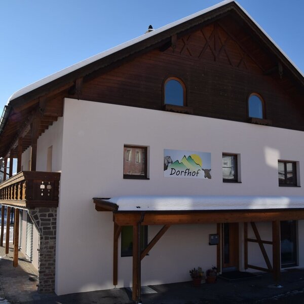 The Dorfhof farmhouse exterior in winter, featuring a wooden balcony, a covered entrance, and the property sign, with snow on the ground and roof.