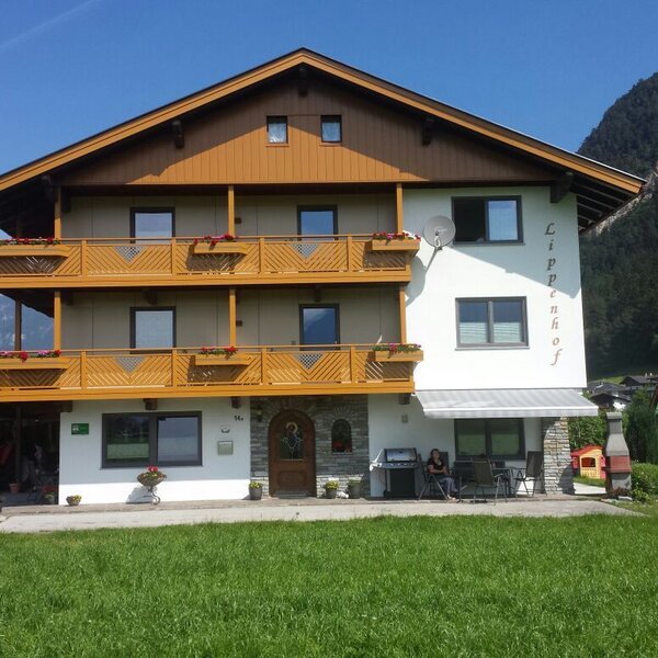 The Farm House exterior, featuring wooden balconies, a ground-floor patio with seating and a barbecue, and a children's playground on the lawn, set against a mountain backdrop.