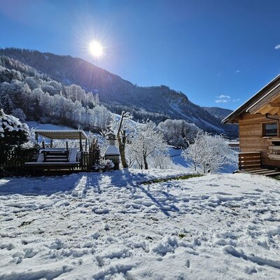 The wooden farmhouse exterior is surrounded by a snow-covered landscape, featuring an outdoor seating area with a pergola and views of the snowy mountains.