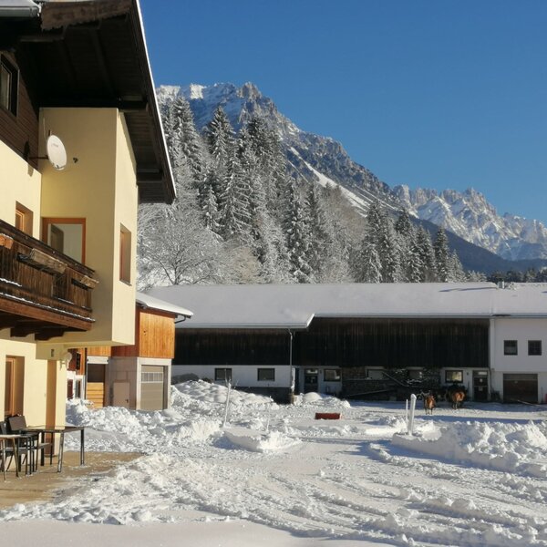 The farm house features a balcony and an outdoor patio with seating, set in a snowy landscape with mountains in the background.