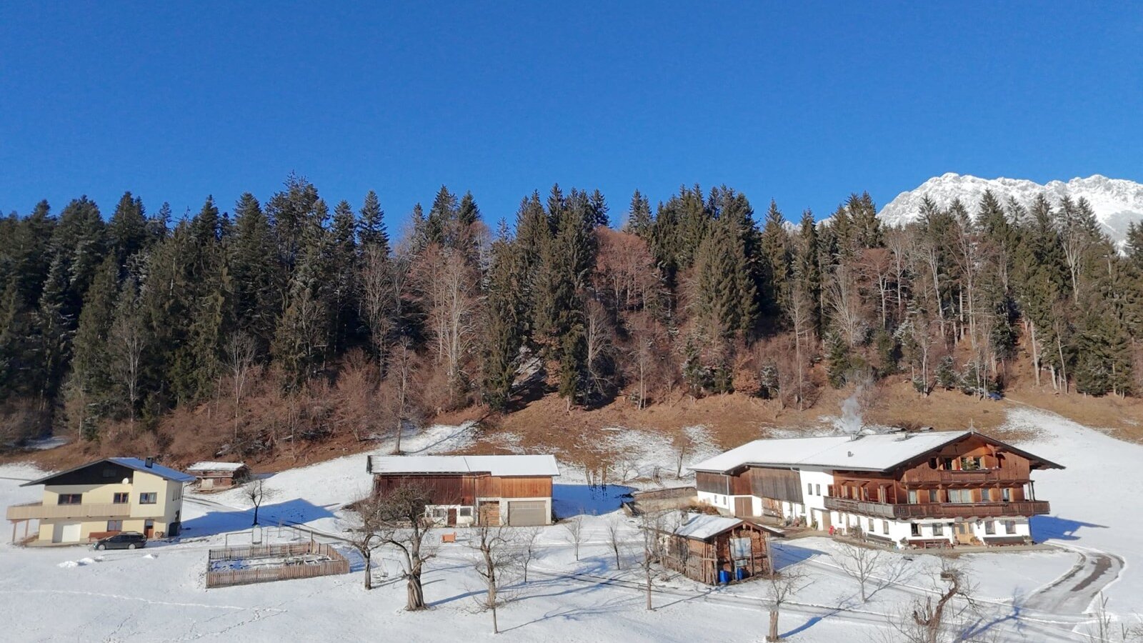 The farmhouse in winter, surrounded by snow, forest, and mountains.