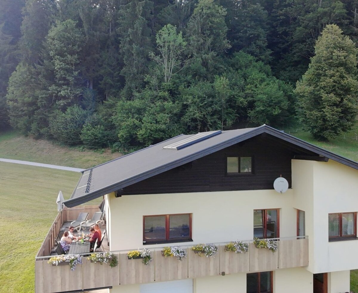 The inviting farmhouse featuring a large, flower-adorned balcony, surrounded by lush green meadows and forests.