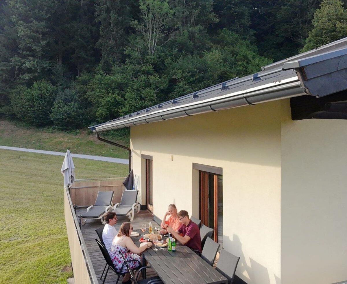 The farmhouse's sunny terrace featuring a large dining table and sun loungers against an idyllic forest backdrop.