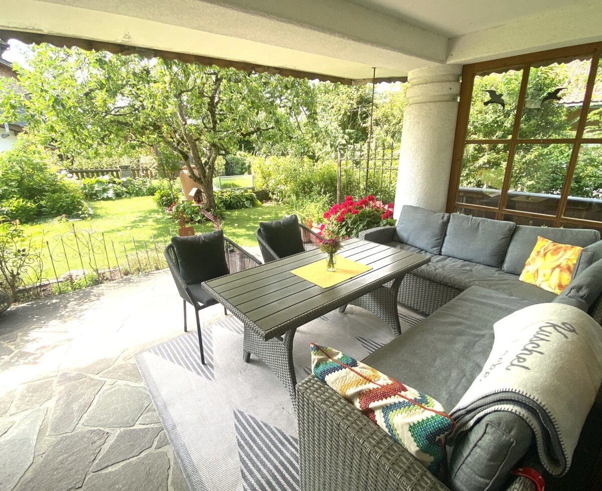The farmhouse's inviting covered terrace, featuring a comfortable lounge area, a dining table, and views of the lush green garden.