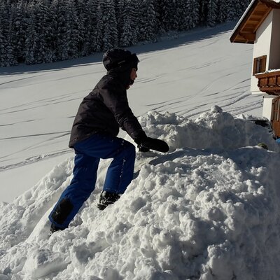 A child playing in a snow pile outside the Alpine Hut, with a snowy field and forest in the background.