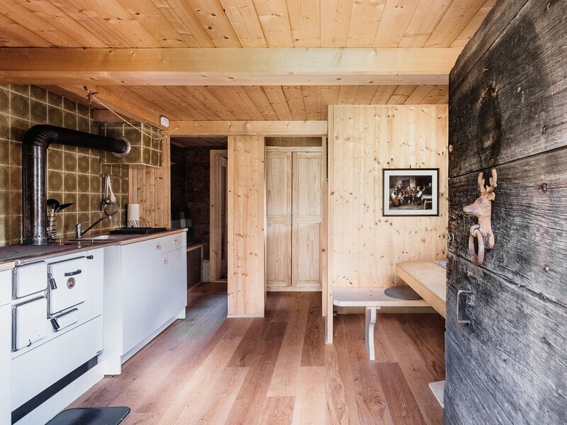 The Alpine Hut features a kitchen equipped with a traditional range cooker, sink, and white cabinets, adjacent to a dining area with a wooden bench and table.
