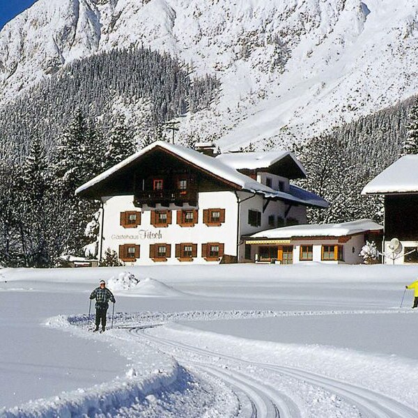 The Farm House with traditional architecture in a snowy landscape, featuring cross-country ski tracks and skiers on the Mieming Plateau.