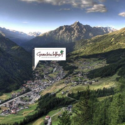 The panoramic mountain valley setting of the farmhouse, featuring a village nestled among forested slopes and peaks.