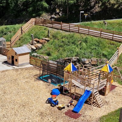 The children's playground at the Farm House includes a wooden climbing frame with a slide, a trampoline, and toy vehicles, set on a wood chip surface.